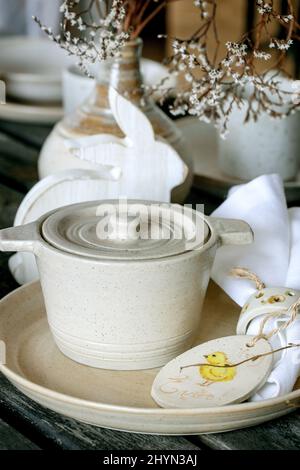 Rustic Easter table setting with empty craft ceramic tableware, cooking pot, plates and bowls, Easter bunny rabbit decorations, vase on wooden table. Stock Photo