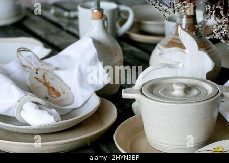 Rustic Easter table setting with empty craft ceramic tableware, cooking pot, plates and bowls, Easter bunny rabbit decorations, vase on wooden table. Stock Photo