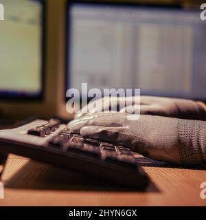 Male hands in dark gloves typing on a computer keyboard against the backdrop of a night office Stock Photo