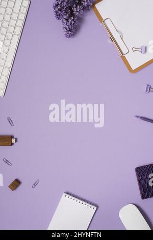 Women's desktop close-up, keyboard, notebooks with pen, lilac flowers. Minimal cropped flat composition on a purple background. Copy space Stock Photo