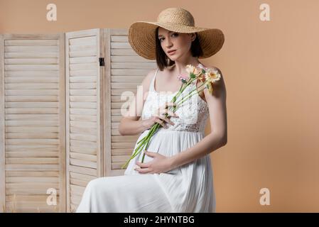 Pregnant woman in dress holding flowers near folding screen isolated on beige Stock Photo