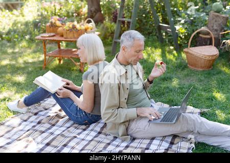 Modern elderly spouses resting in garden, man using laptop computer while his wife reading book, sitting back to back Stock Photo
