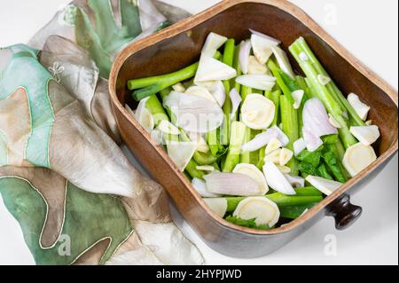 Rustic earthenware baking pan with pile of sliced raw vegetable, preparation for baking, green scarf on white background. Many spring vegetable in pan Stock Photo
