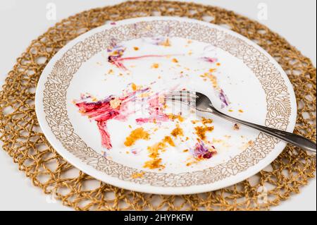 Dirty empty plate with crumbs from puff pastry and colorful fleck from fruit, fork, bamboo pad on white background. Stock Photo