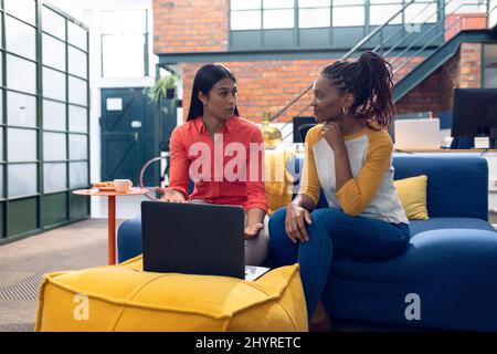 Multiracial young businesswomen discussing over laptop while sitting on couch in modern office Stock Photo