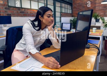 Biracial young businesswoman with hand on chin working on laptop while sitting at desk in office Stock Photo