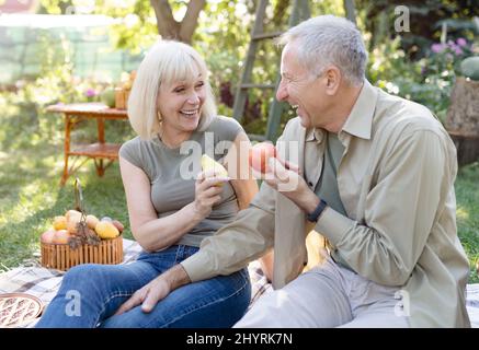 Laughing senior spouses sitting on field grass and eating fruits, talking and smiling, resting outdoors in their garden Stock Photo