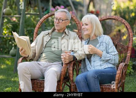 Loving elderly couple reading book together, sitting in wicker chairs and spending spring evening in their garden Stock Photo