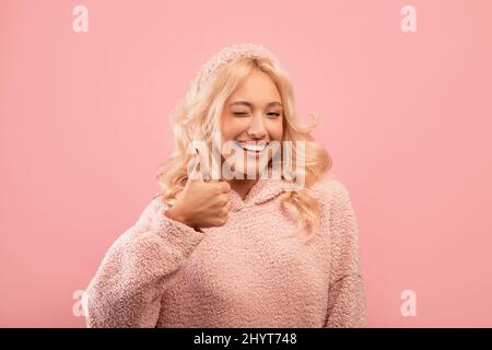 Pretty stylish lady showing thumb up, winking and smiling at camera, recommending something, pink background Stock Photo