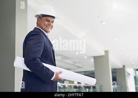 I am officially ready to begin. Portrait of a cheerful professional male architect looking at the camera while holding blueprints inside a building. Stock Photo