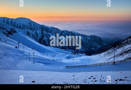 Beautiful landscape Shymbulak mountain resort snowy empty ski slope and cable car lift at sunset orange sky in Almaty, Kazakhstan. Stock Photo
