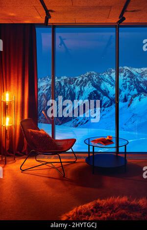 Loft Interior with chair and table in the room near panoramic window with blue mountain and glow orange lamp in hotel at Shymbulak ski resort Almaty, Stock Photo