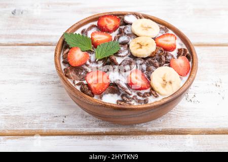 Chocolate cornflakes with milk and strawberry in wooden bowl on white wooden background. Side view, close up. Stock Photo