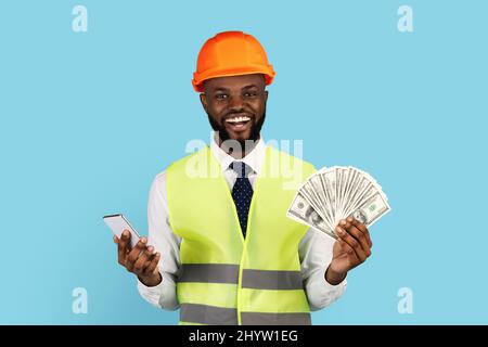 Happy Construction Worker With Money Cash And Smartphone Posing Over Blue Background Stock Photo