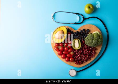 Overhead view of healthy food on heart shaped cutting board by stethoscope on blue background Stock Photo