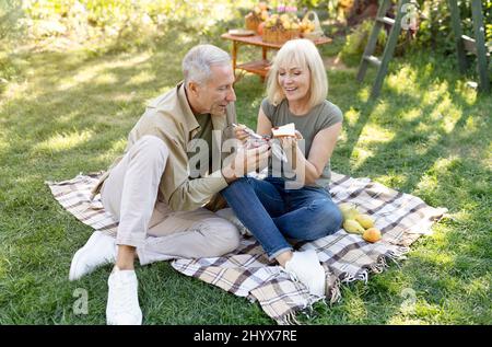 Cute retired couple having picnic in garden, sitting on blanket and eating toasts with jam, enjoying warm spring day Stock Photo