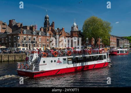 City Cruises boat sailing on water & people sitting outside cafes - busy sunny picturesque River Ouse, King's Staith, York, North Yorkshire, England.