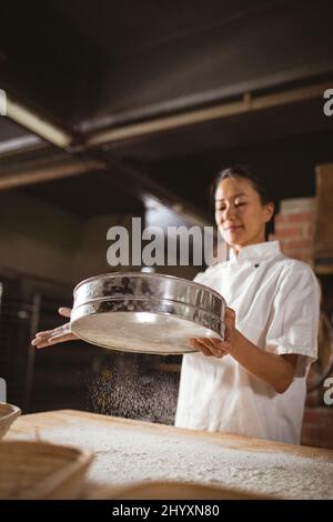 Asian mid adult female baker straining flour on table while working in bakery Stock Photo