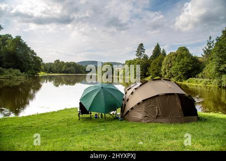 A tent and large umbrella stand on the bank of a lake surrounded by beautiful trees. A peaceful location to relax and enjoy the great outdoors. Stock Photo