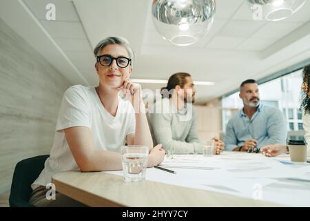 Female design professional looking at the camera while sitting in a meeting with her team. Group of creative businesspeople brainstorming while workin Stock Photo