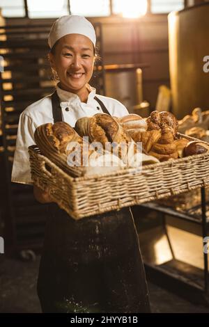 Portrait of smiling asian mid adult female baker holding loaf of breads basket in basket Stock Photo
