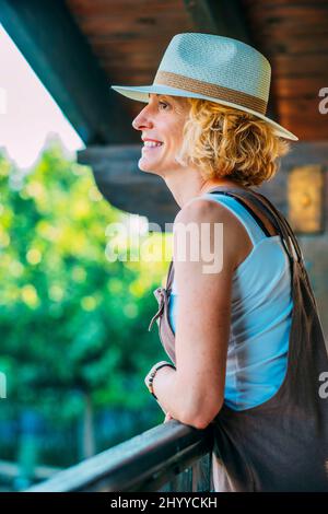 Portrait of a mature caucasian blonde young woman in her 50s wearing a hat  on the wooden porch of a country house. Lifestyle concept. Stock Photo