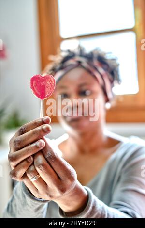 Closeup portrait of a young black woman with afro hairstyle joking with a lollipop indoor at home. Lifestyle concept. Stock Photo
