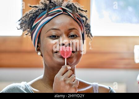 Closeup funny portrait of a young black woman with afro hairstyle joking with a lollipop indoor at home, laughing. Lifestyle concept. Stock Photo