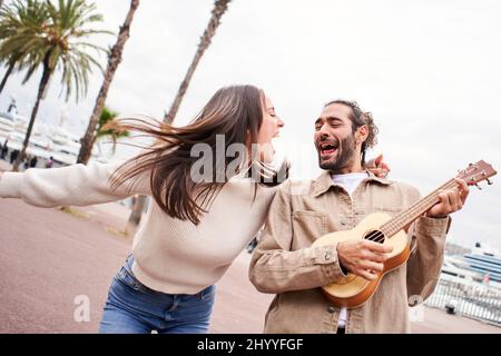 Funny happy and loving couple dance outdoors on street singing and playing guitar. Man and woman have fun during their holiday at city Stock Photo