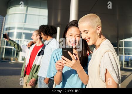 Group of happy friends using the mobile in the street. Young smiling people sharing with cell phone outdoors. Stock Photo