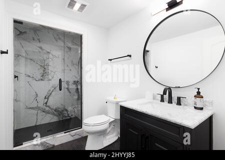 A beautiful bathroom with a dark wood vanity cabinet, large circular mirror, small herringbone tile flooring, and a walk-in marble tiled shower. Stock Photo