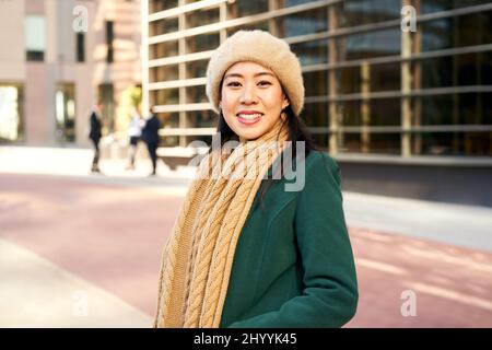 An Asian woman looking at the camera outside an office area with business people in the background. Cheerful Chinese girl in winter clothes in the Stock Photo