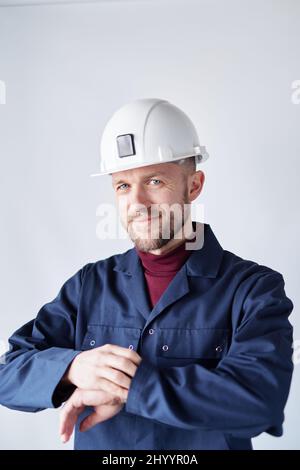Construction industry concept. Attractive bearded caucasian engineer, worker, supervisor or building contractor in white helmet and blue uniform looking checking time. High quality vertical image Stock Photo