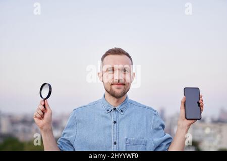 Information searching concept. Male wearing jeans shirt holding magnifying glass and smartphone in hands at city view as detective or investigator. Young bearded man happy expression looking at camera Stock Photo