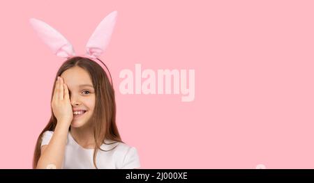 Picture of a blond little girl wearing bunny ears on head smiling for the camera and covering one eye with a palm on pink background and copy space. Stock Photo