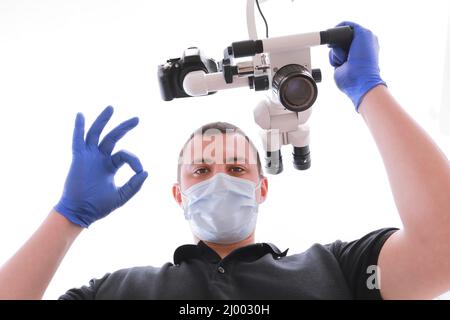 male dentist in uniform, medical mask and glovers looking at camera and showing fingers ok. Young doctor sitting working in dental office with modern dental microscope. Concept of stomatology. Stock Photo