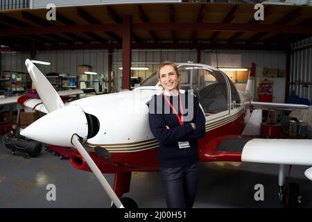 Young female pilot posing smiling in front of her plane inside the hangar. Stock Photo