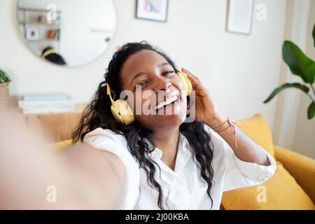 Happy selfie of cheerful young woman listening to music with headphones on soft couch at home in day off. Stock Photo