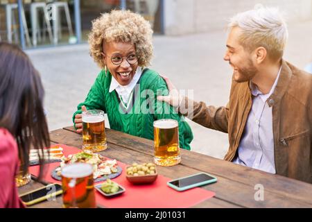 Multiracial couple having fun. Happy people celebrating drinking beer together with friends at terrace bar. Man and woman laughing outdoors. Stock Photo