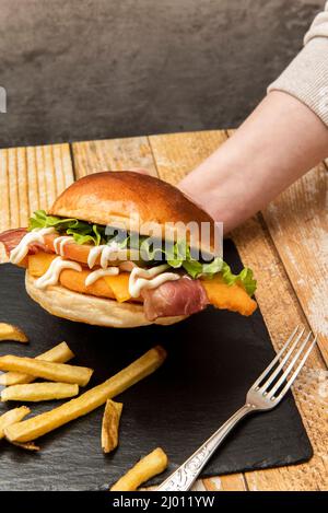 A hand tightly holding a crispy chicken burger with freshly cut tomatoes, fresh lettuce and mayonnaise on a black slate plate with french fries and a Stock Photo