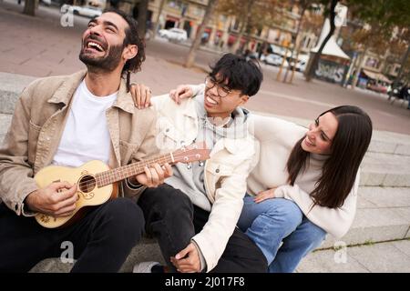 Happy people singing outdoors sitting in street. Group of friends having fun playing the ukulele. Stock Photo