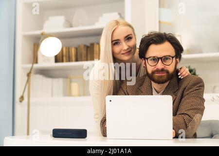 Young Married Caucasian Couple portrait sitting in front of the laptop smiling to the camera hugging inside / Remote work concept . High quality photo Stock Photo
