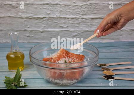 Salting salmon at home,  fillet in a transparent bowl olive oil and various spices in wooden spoons on the table Stock Photo
