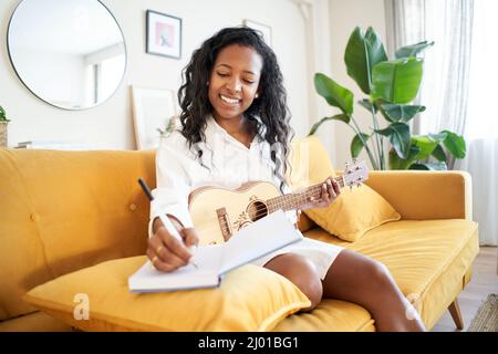 Woman singer-songwriter composing a song at home. Musical artist creating. Girl writing a song in a notebook sitting on the sofa. Stock Photo