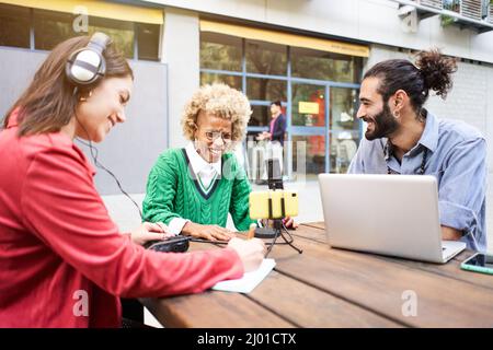 Young woman interviewing a man in a casual radio studio outdoors. Millennial people using technology, computers, headphones, microphone... Stock Photo