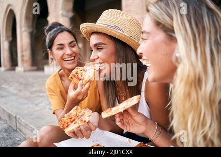 Three beautiful women sitting on the stairs of the city streets eating pizza in a street stall. The happy girls enjoy the weekend together. Stock Photo