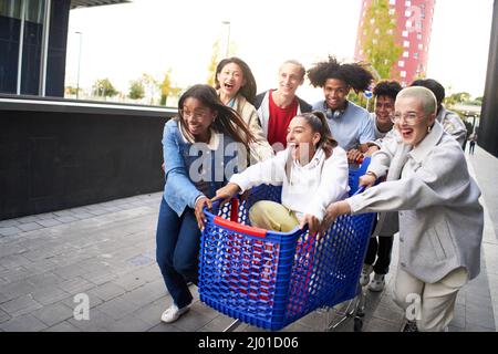Young multiracial people having fun together with shopping cart. Millennial friends sharing time with trolleys at commercial mall parking. Youth Stock Photo
