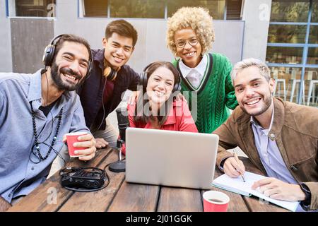 Portrait looking at the camera of a Group of young smiling people outdoors watching a video on the laptop. Colleagues broadcasting togethers Stock Photo