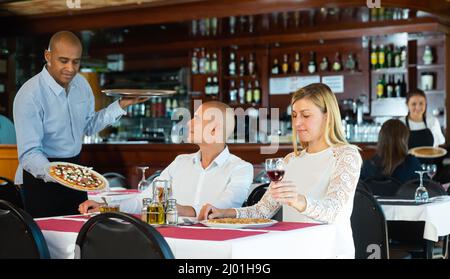 Smiling hispanic waiter bringing pizzas to couple in pizzeria Stock Photo