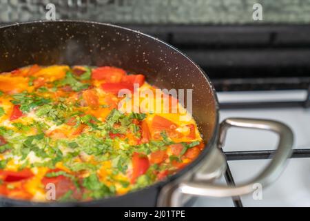 Fried eggs with vegetables and herbs in black frying pan on white stove. Side view Stock Photo